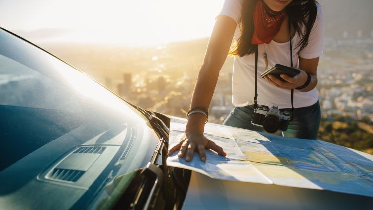 Solo travel safety apps are essential on a road trip, as shown by this woman with a map open on her car and a smartphone in her hand.