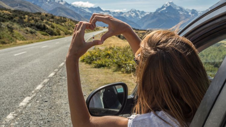 woman leaning out of her car window making a heart shape with her hands indicating that she is loving being on a solo road trip