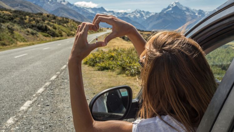 mulher inclinando-se para fora da janela do carro fazendo um coração com as mãos, indicando que ela está amando estar em uma viagem solo